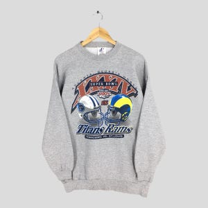 Puede incluir: Una sudadera gris con un gráfico del Super Bowl XXXIV. El diseño incluye cascos de fútbol de los Tennessee Titans y los St. Louis Rams. El texto dice "Atlanta Georgia Hosts" y "Titans Rams Tennessee vs. St. Louis".