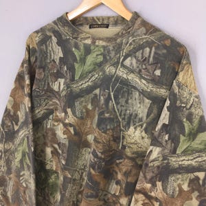 K&ouml;nnte beinhalten: Ein Camouflage-Sweatshirt mit Rundhalsausschnitt und langen &Auml;rmeln, das ein Muster aus braunen, gr&uuml;nen und beigen Bl&auml;ttern und &Auml;sten zeigt. Auf dem Kragen steht "COMMANDER". Das Muster ist "Advantage Timber".