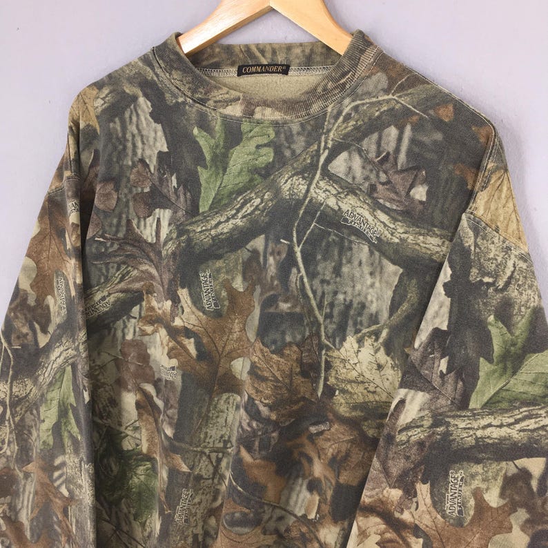 K&ouml;nnte beinhalten: Ein Camouflage-Sweatshirt mit Rundhalsausschnitt und langen &Auml;rmeln, das ein Muster aus braunen, gr&uuml;nen und beigen Bl&auml;ttern und &Auml;sten zeigt. Auf dem Kragen steht "COMMANDER". Das Muster ist "Advantage Timber".