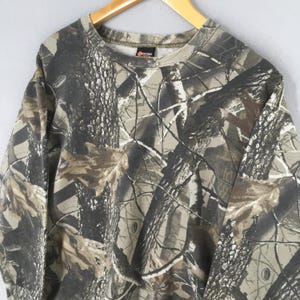 May include: A long-sleeved sweatshirt with a camouflage pattern in shades of brown, green, and gray. The sweatshirt has a crew neck and a tag on the collar. The fabric appears to be a medium weight material.