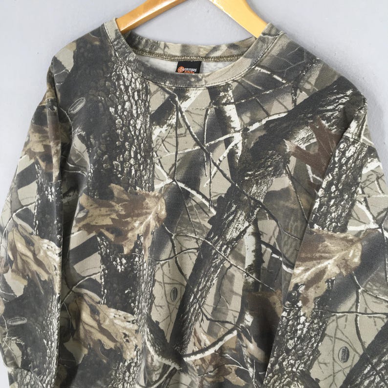 May include: A long-sleeved sweatshirt with a camouflage pattern in shades of brown, green, and gray. The sweatshirt has a crew neck and a tag on the collar. The fabric appears to be a medium weight material.