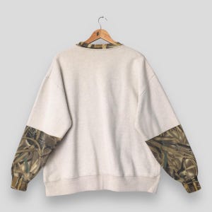 May include: A light beige sweatshirt with camouflage patterned sleeves and collar. The sleeves and collar feature a brown, green, and tan camouflage design. The sweatshirt is hanging on a wooden hanger.