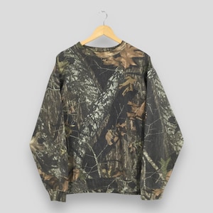 Puede incluir: Una sudadera con estampado de camuflaje en tonos marrones, verdes y negros, colgada de una percha de madera. La parte superior de manga larga presenta cuello redondo y dobladillos y pu&ntilde;os acanalados. La tela tiene un estampado denso y detallado que se asemeja a un bosque.