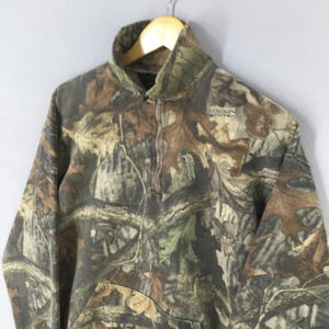 May include: A camouflage-patterned fleece pullover with a high collar and a quarter-zip closure. The fabric features a mix of brown, green, and tan hues, mimicking a forest environment. The brand name "Advantage" is visible on the garment.