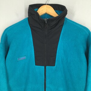 May include: A teal fleece jacket with a black collar and zipper. The jacket has a high collar and a small logo patch on the left chest. The jacket is hanging on a wooden hanger.