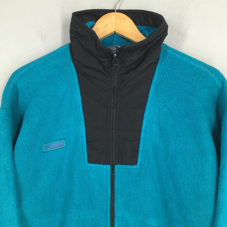 May include: A teal fleece jacket with a black collar and zipper. The jacket has a high collar and a small logo patch on the left chest. The jacket is hanging on a wooden hanger.