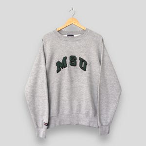 Puede incluir: Una sudadera gris con las letras "MSU" en verde oscuro, arqueadas sobre el pecho. La sudadera tiene cuello redondo y mangas largas. La prenda está colgada de una percha de madera.
