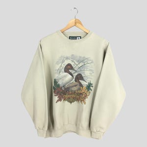 Peut inclure: Un sweat-shirt beige avec un imprim&eacute; graphique de deux canards dans un cadre naturel. Les canards sont bruns et blancs avec une illustration d&eacute;taill&eacute;e de leurs plumes. Le texte "Snowshoe" est imprim&eacute; sur le sweat-shirt.