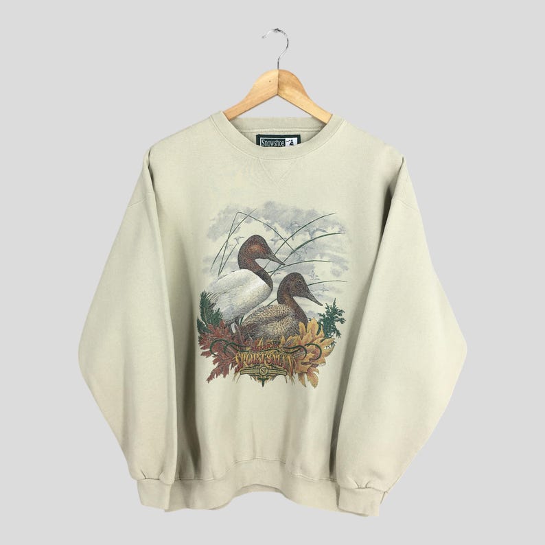 Peut inclure: Un sweat-shirt beige avec un imprim&eacute; graphique de deux canards dans un cadre naturel. Les canards sont bruns et blancs avec une illustration d&eacute;taill&eacute;e de leurs plumes. Le texte "Snowshoe" est imprim&eacute; sur le sweat-shirt.