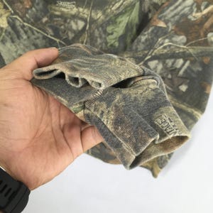 May include: Close-up of a camouflage-patterned long-sleeve shirt. The fabric features a mix of green, brown, and tan hues. The cuff of the sleeve is visible, with a ribbed texture and a tag with the word "VINTAGE".