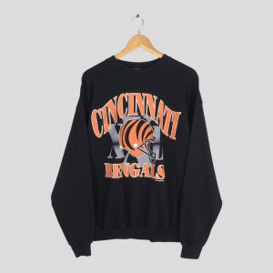 May include: A black long-sleeve sweatshirt featuring a graphic print of a football helmet with orange and black tiger stripes. The text reads CINCINNATI BENGALS in orange lettering, with a large grey XL logo positioned behind the helmet graphic.