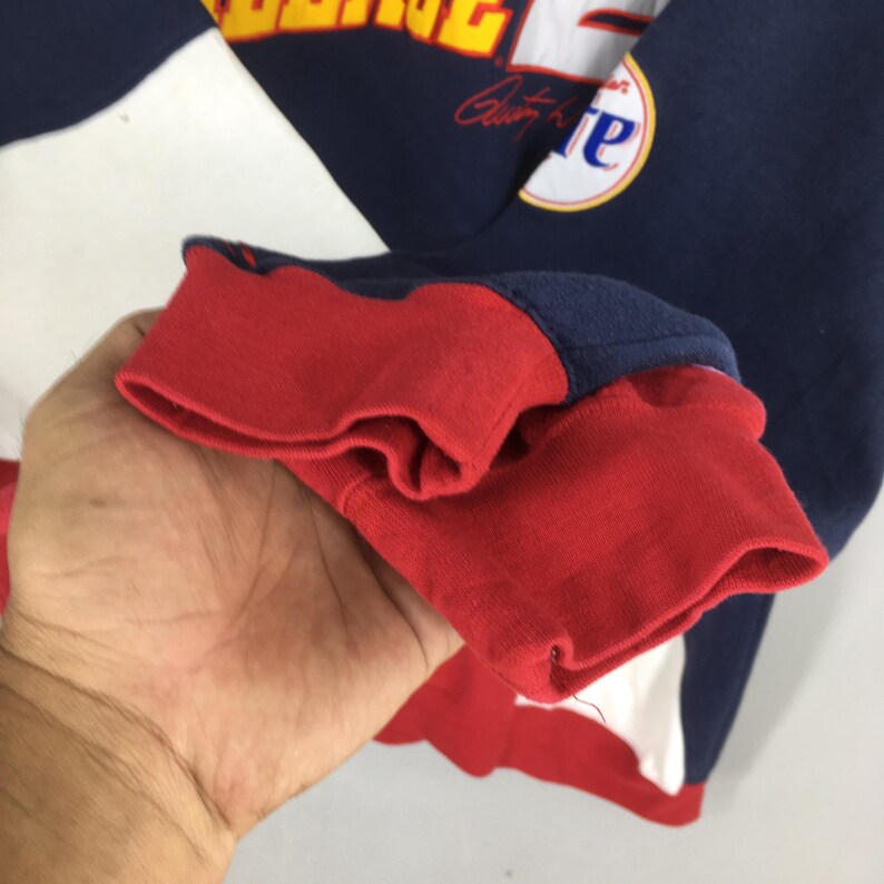May include: A close-up of a navy blue and red sweatshirt sleeve. The sleeve cuff is red, and the upper sleeve is navy blue with white and yellow lettering. A circular patch with the letter "e" is visible.