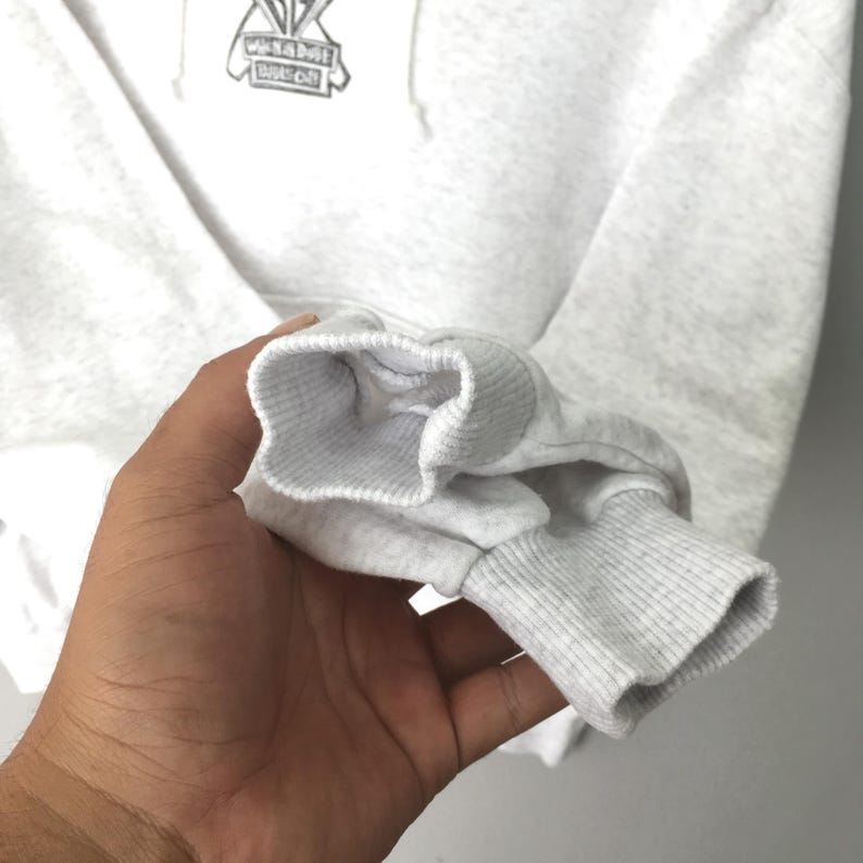 Puede incluir: Primer plano de una manga de sudadera con capucha gris claro con pu&ntilde;o acanalado. La sudadera tiene un gr&aacute;fico blanco en la parte superior del pecho. La manga est&aacute; sujeta por una mano.