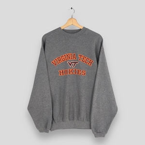 Może przedstawiać: Szara bluza z okrągłym dekoltem z napisem "VIRGINIA TECH" w kolorze pomarańczowym i kasztanowym na piersi. Poniżej znajduje się napis "VT HOKIES" w tych samych kolorach. Bluza wisi na drewnianym wieszaku.