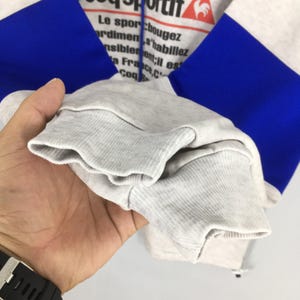 May include: A close-up of a gray, blue, and white hoodie. The hoodie has blue sleeves and a gray body with text at the top. The cuff of the sleeve is gray and ribbed.