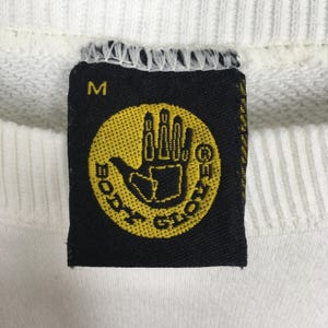Op de afbeelding: Close-up van een witte sweatshirt met een zwart label. Het label heeft een geel cirkelvormig logo met een handontwerp en de woorden "BODY GLOVE". Het label heeft ook de letter "M".