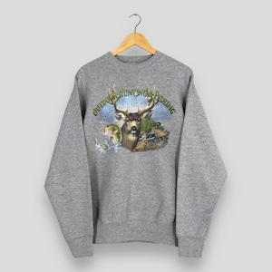 Puede incluir: Una sudadera gris con un gráfico que presenta un ciervo, un pez y un pato, con el texto "Outdoor Hunting & Fishing" en un diseño circular. La imagen tiene un tema de naturaleza.