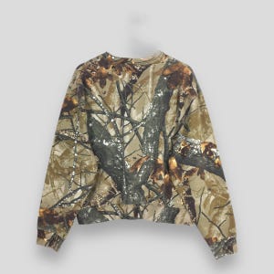 May include: A camouflage-patterned sweatshirt in shades of brown, green, and tan. The long-sleeved top features a crew neck and a dense forest design, ideal for outdoor activities or casual wear. The fabric appears to be a medium weight.