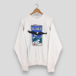 May include: White sweatshirt featuring a graphic of an eagle with outstretched wings, the word "ALASKA", a helicopter, and mountain scenery. The design is in a blue and black rectangular frame. The sweatshirt is hanging on a wooden hanger.