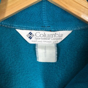 May include: Close-up of a teal fleece garment with the Columbia Sportswear Company label. The label is white with the brand name and logo, and the text "PORTLAND, OREGON U.S.A."