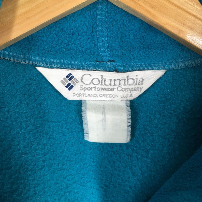 May include: Close-up of a teal fleece garment with the Columbia Sportswear Company label. The label is white with the brand name and logo, and the text "PORTLAND, OREGON U.S.A."