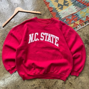 Puede incluir: Una sudadera roja de cuello redondo con el texto "N.C. STATE" en letras blancas. La sudadera está colgada en una percha de madera. En el fondo se ve una alfombra colorida.
