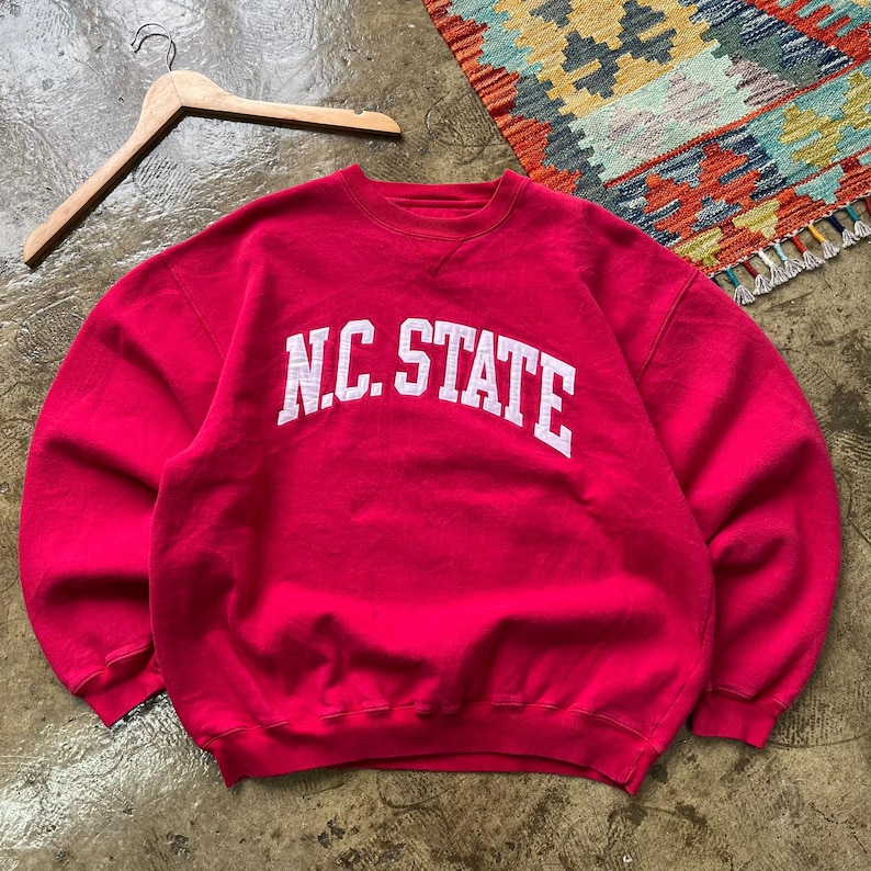 May include: A red crewneck sweatshirt with the text "N.C. STATE" in white block letters. The sweatshirt is hanging on a wooden hanger. The background includes a colorful rug.