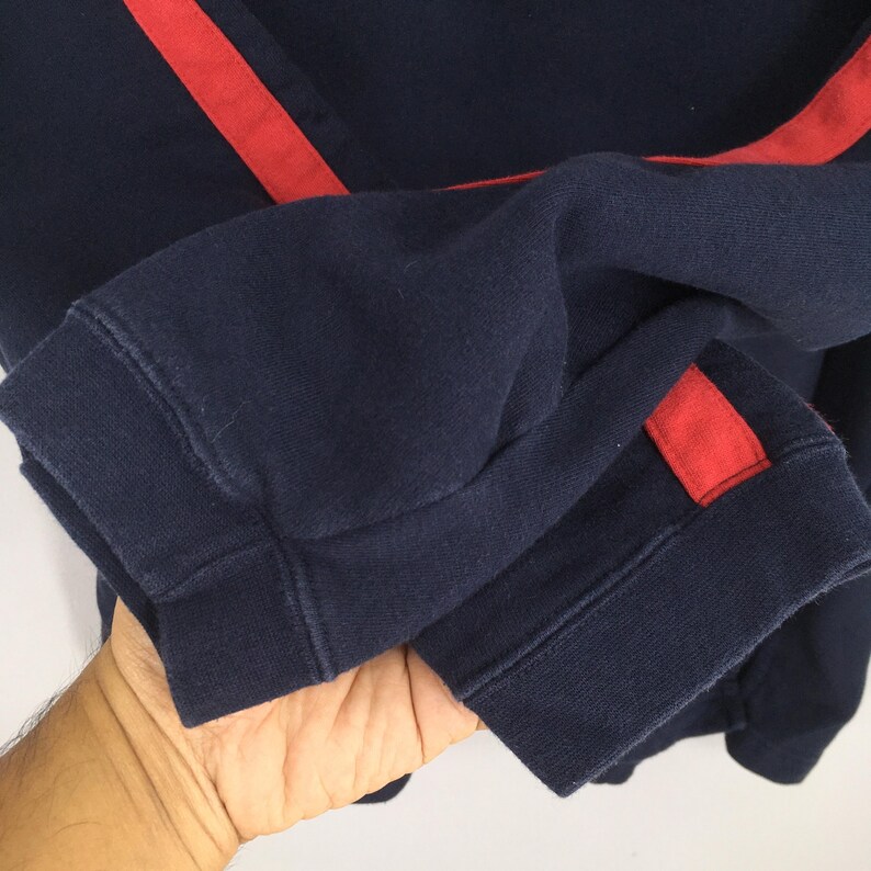 Puede incluir: Primer plano de una sudadera azul marino con un detalle de raya roja. Los pu&ntilde;os y el dobladillo son acanalados. La tela parece ser un material suave y c&oacute;modo. La raya roja recorre la manga.