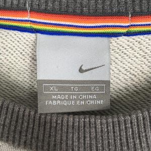 K&ouml;nnte beinhalten: Nahaufnahme eines grauen Nike Sweatshirt-Etiketts. Das Etikett zeigt das Nike-Logo und den Text "MADE IN CHINA FABRIQUE EN CHINE". Ein Regenbogenstreifen-Detail ist oben auf dem Sweatshirt zu sehen.