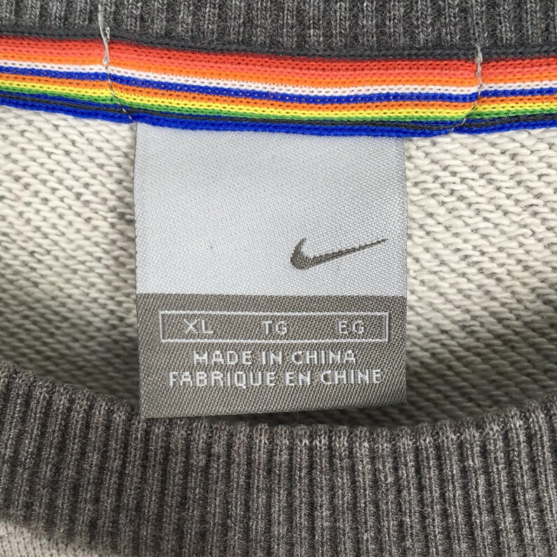 K&ouml;nnte beinhalten: Nahaufnahme eines grauen Nike Sweatshirt-Etiketts. Das Etikett zeigt das Nike-Logo und den Text "MADE IN CHINA FABRIQUE EN CHINE". Ein Regenbogenstreifen-Detail ist oben auf dem Sweatshirt zu sehen.