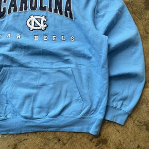 K&ouml;nnte beinhalten: Hellblaues Kapuzen-Sweatshirt mit dem Wort "CAROLINA" in Marineblau oben. Darunter befindet sich ein wei&szlig;es und marineblaues Logo. Das Wort "HEELS" ist in Marineblau gedruckt. Das Sweatshirt hat eine Vordertasche.