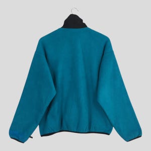 May include: Teal fleece jacket with a black collar and trim, hanging on a hanger. The jacket has a high collar and long sleeves. The back of the jacket is visible, showcasing the teal color and the black accents.