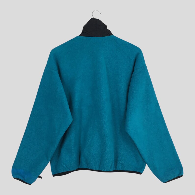 May include: Teal fleece jacket with a black collar and trim, hanging on a hanger. The jacket has a high collar and long sleeves. The back of the jacket is visible, showcasing the teal color and the black accents.