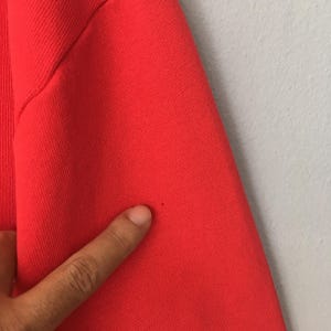 Peut inclure: Un v&ecirc;tement en tissu rouge vif pr&eacute;sentant une texture c&ocirc;tel&eacute;e le long de la couture lat&eacute;rale. Le mat&eacute;riau affiche une petite tache sombre ou une imperfection unique sur la surface, soulignant la texture et la couleur du textile en gros plan.