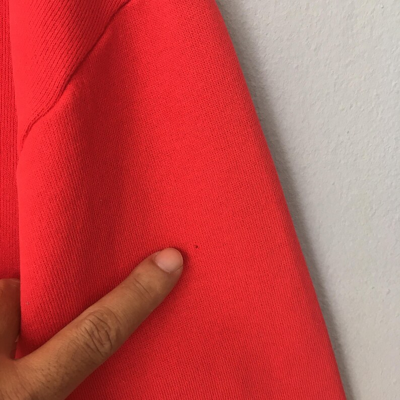 Peut inclure: Un v&ecirc;tement en tissu rouge vif pr&eacute;sentant une texture c&ocirc;tel&eacute;e le long de la couture lat&eacute;rale. Le mat&eacute;riau affiche une petite tache sombre ou une imperfection unique sur la surface, soulignant la texture et la couleur du textile en gros plan.