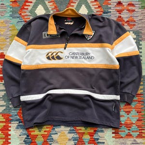 May include: A navy blue long-sleeve rugby shirt with white and gold accents. The shirt features a zip-up collar and the text "Canterbury of New Zealand" across the chest. The shirt has a classic rugby design.