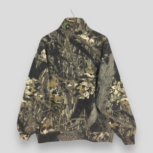 May include: A camouflage-patterned zip-up sweatshirt. The jacket features a high collar and a design of brown, green, and tan foliage. The sweatshirt is hanging on a metal hanger against a light gray background.