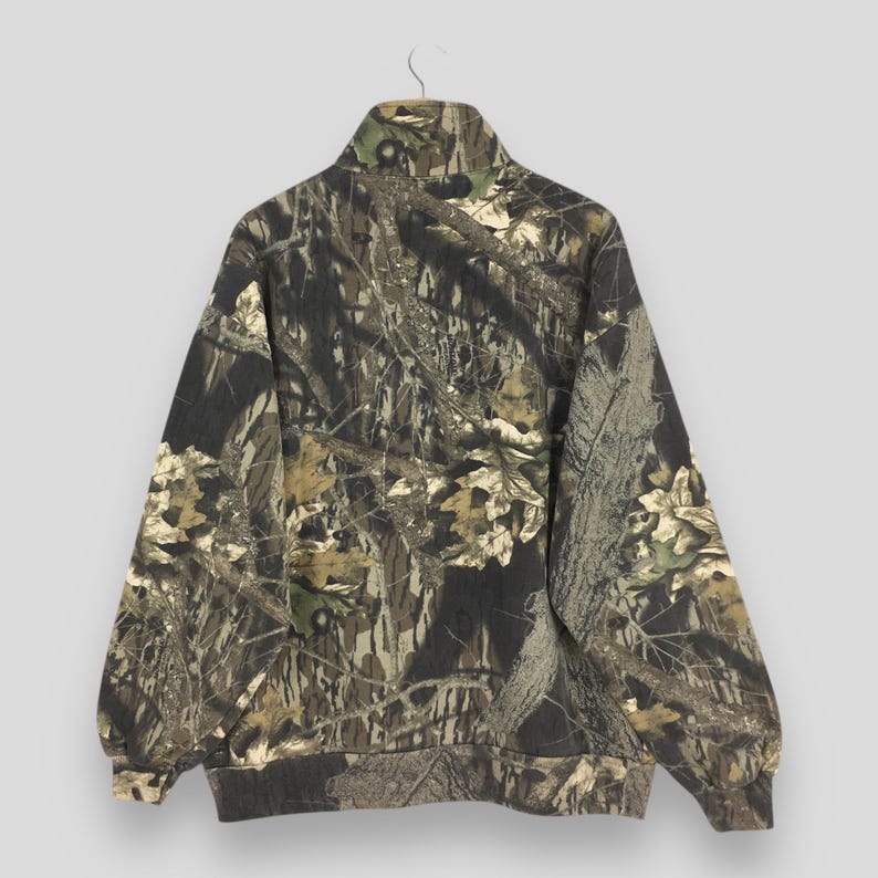 May include: A camouflage-patterned zip-up sweatshirt. The jacket features a high collar and a design of brown, green, and tan foliage. The sweatshirt is hanging on a metal hanger against a light gray background.