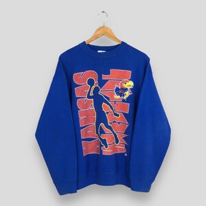 May include: A blue sweatshirt with a red and white graphic that says "Kansas" with a basketball player silhouette. The sweatshirt also features a "KU" logo with a Jayhawk mascot.