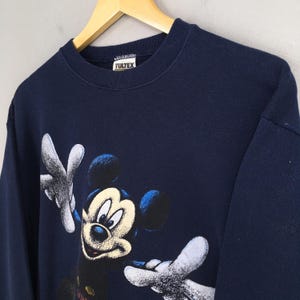 May include: Navy blue sweatshirt featuring a graphic of Mickey Mouse with outstretched arms. The cartoon character is in a classic pose with white gloves and a smiling face. The sweatshirt has a crew neck and the brand name "YULTEX" is visible on the tag.