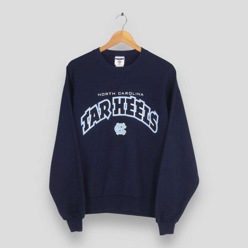 May include: Navy blue sweatshirt with the text "NORTH CAROLINA TAR HEELS" in white and light blue. The sweatshirt has a crew neck and long sleeves. A light blue logo is below the text.