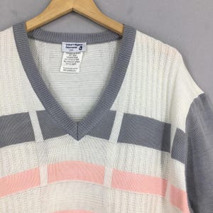 May include: A vintage V-neck sweater in white, gray, and pink. The sweater features a geometric design with gray sleeves and trim. The label reads "courr&egrave;ges homme".
