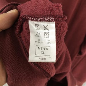 May include: Close-up of a burgundy garment tag. The tag shows care symbols, fabric composition percentages, and the text "MEN'S XL". The garment appears to be made of a soft, textured material.