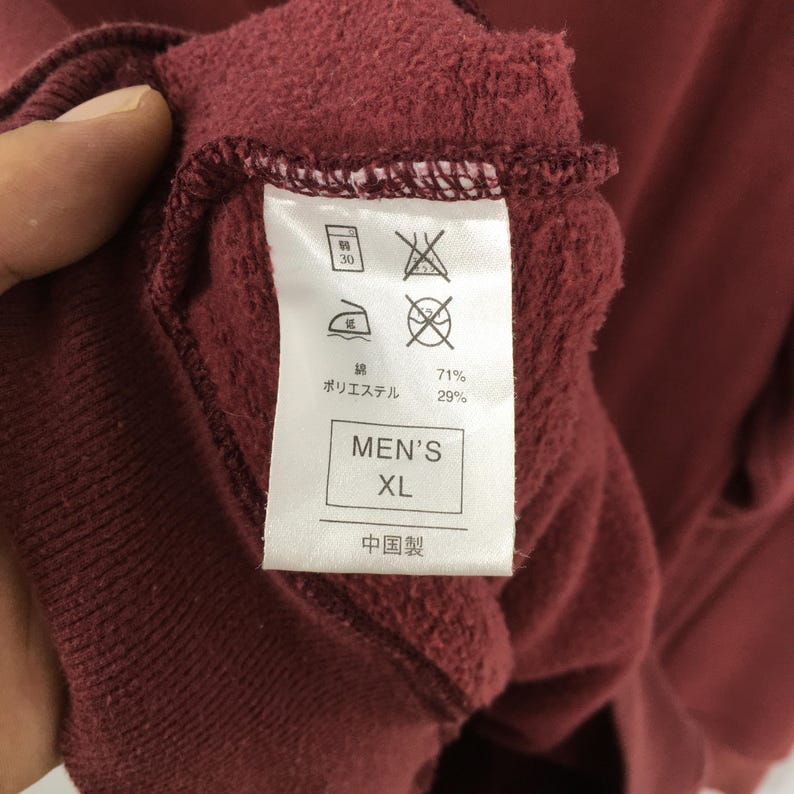 May include: Close-up of a burgundy garment tag. The tag shows care symbols, fabric composition percentages, and the text "MEN'S XL". The garment appears to be made of a soft, textured material.