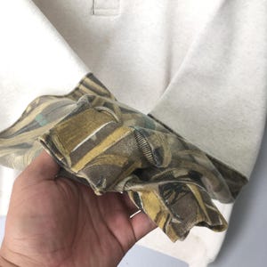 May include: A cream-colored long-sleeve shirt with a camouflage pattern on the cuffs. The cuffs feature a mix of brown, green, and tan hues. The shirt has a simple, classic design.