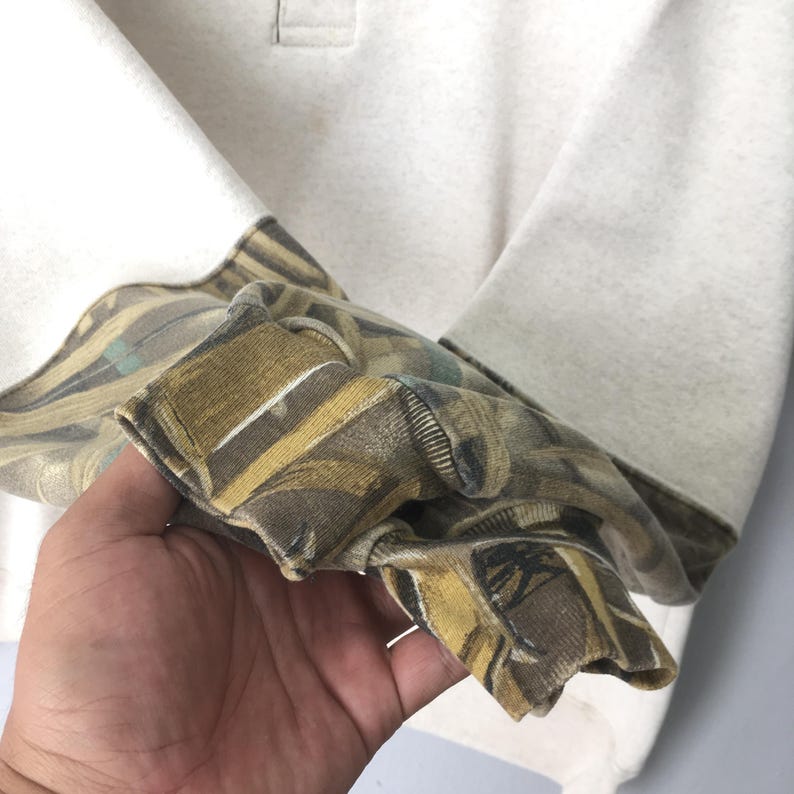 May include: A cream-colored long-sleeve shirt with a camouflage pattern on the cuffs. The cuffs feature a mix of brown, green, and tan hues. The shirt has a simple, classic design.