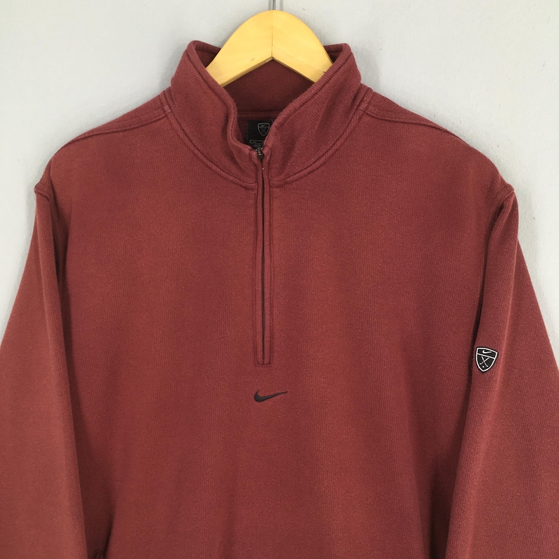 May include: A maroon sweatshirt with a quarter-zip and a high collar. The Nike logo is on the chest, and a shield logo is on the left sleeve. The sweatshirt is displayed on a wooden hanger.