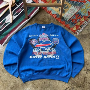 以下が含まれることがあります： Buffalo Billsのグラフィックが施された青いクルーネックのスウェットシャツ。Buffalo Bills、Super Bowl XXVI、XXVII、XXV、1992 AFC Champions、Sweet Repeatの文字がプリントされています。デザインにはチームロゴと様々なスーパーボウルのエンブレムが含まれています。