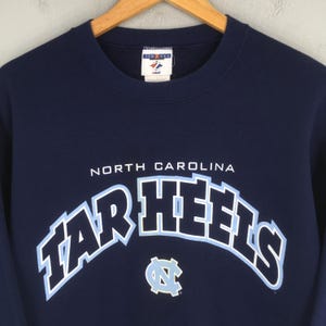 May include: Navy blue sweatshirt with the words "NORTH CAROLINA" above the arched "TAR HEELS" in light blue and white. A light blue and white logo is below the text. The sweatshirt is hanging on a wooden hanger.