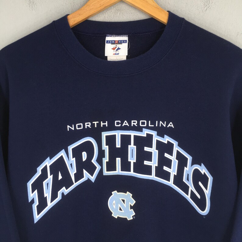 May include: Navy blue sweatshirt with the words "NORTH CAROLINA" above the arched "TAR HEELS" in light blue and white. A light blue and white logo is below the text. The sweatshirt is hanging on a wooden hanger.
